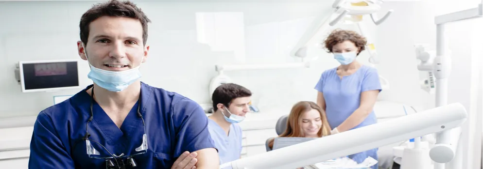 bandeau-dentistes1 Praticien souriant en tenue bleue, bras croisés, et équipe soignante au fauteuil d'un cabinet dentaire