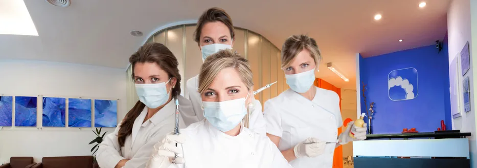 bandeau-groupe-assistante1 Quatre soignantes en blouse blanche et masque posant ensemble dans un cabinet aux murs bleus